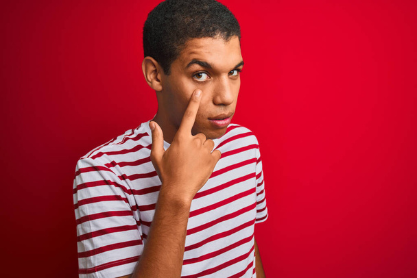 Joven hombre árabe guapo usando una camiseta a rayas sobre un fondo rojo aislado señalando al ojo observándote gesto, expresión sospechosa
 - Foto, Imagen
