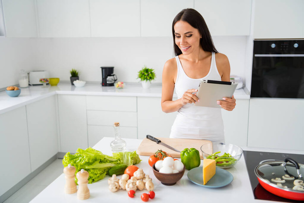 Portrait of beautiful positive brown haired woman want cook tasty yummy healthcare dinner use tablet find recipe follow look at useful fresh ingredients wear white singlet in house kitchen - Фото, изображение