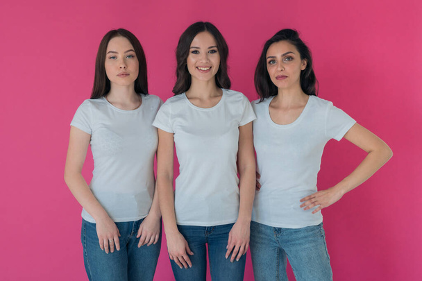 Three beautiful brunettes in white t-shirts and jeans on a pink background. Logos on clothes. T-shirt design - Fotoğraf, Görsel
