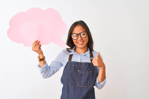 Chinese woman wearing glasses apron holding speech bubble over isolated white background happy with big smile doing ok sign, thumb up with fingers, excellent sign - Valokuva, kuva