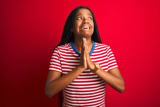 Fiatal afro-amerikai nő csíkos pólóban áll elszigetelt vörös háttér felett, könyörög és imádkozik kéz a kézben, reményteljes arckifejezéssel nagyon érzelmes és aggódó. Bocsánatot kér. Vallási fogalom. - Fotó, kép