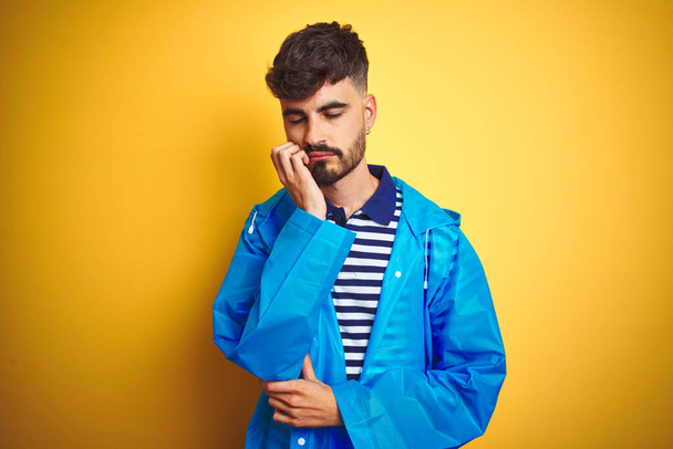 Young handsome man wearing rain coat standing over isolated yellow background thinking looking tired and bored with depression problems with crossed arms. - Photo, image
