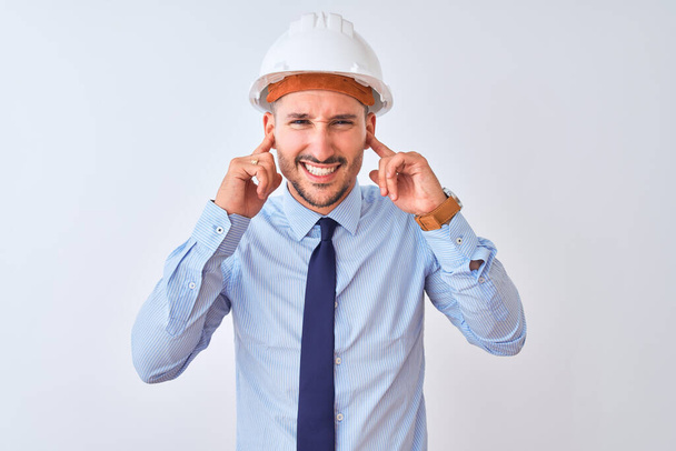 Young business man wearing contractor safety helmet over isolated background covering ears with fingers with annoyed expression for the noise of loud music. Deaf concept. - Fotó, kép