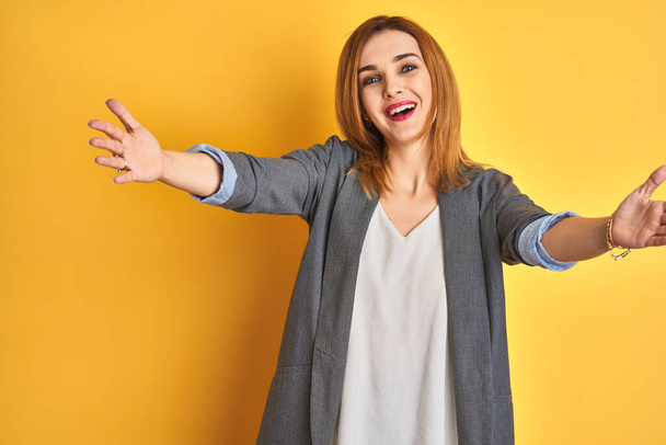 Redhead caucasian business woman over yellow isolated background looking at the camera smiling with open arms for hug. Cheerful expression embracing happiness. - Valokuva, kuva