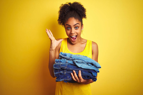 Joven mujer afroamericana sosteniendo pila de jeans sobre aislado fondo amarillo muy feliz y emocionado, expresión ganadora celebrando victoria gritando con gran sonrisa y las manos levantadas
 - Foto, Imagen