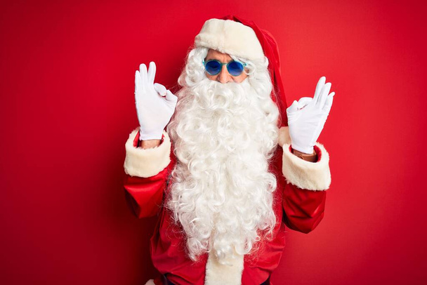 Mężczyzna w średnim wieku w kostiumie Świętego Mikołaja i okularach przeciwsłonecznych na izolowanym czerwonym tle relaks i uśmiech z zamkniętymi oczami robi gest medytacji palcami. Koncepcja jogi. - Zdjęcie, obraz