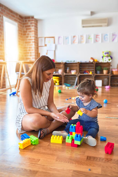 Young beautiful teacher and toddler playing with building blocks toy at kindergarten - Photo, Image
