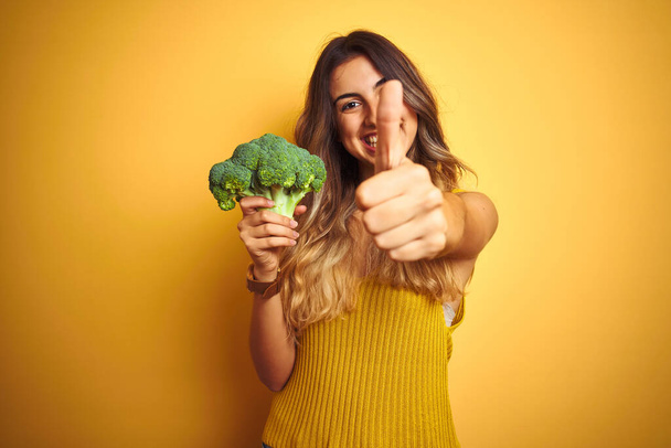 Jovem mulher bonita comendo brócolis sobre amarelo isolado fundo feliz com grande sorriso fazendo ok sinal, polegar para cima com os dedos, excelente sinal
 - Foto, Imagem