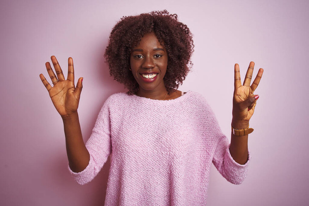 Jonge Afrikaanse vrouw draagt trui die over een geïsoleerde roze achtergrond staat te kijken en te wijzen met vingers nummer acht terwijl ze zelfverzekerd en gelukkig glimlacht. - Foto, afbeelding