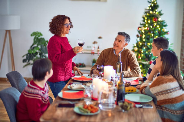 schöne Familie lächelt glücklich und zuversichtlich. einer von ihnen steht mit einem Becher Wein in der Hand und spricht eine Weihnachtsansprache zu Hause - Foto, Bild