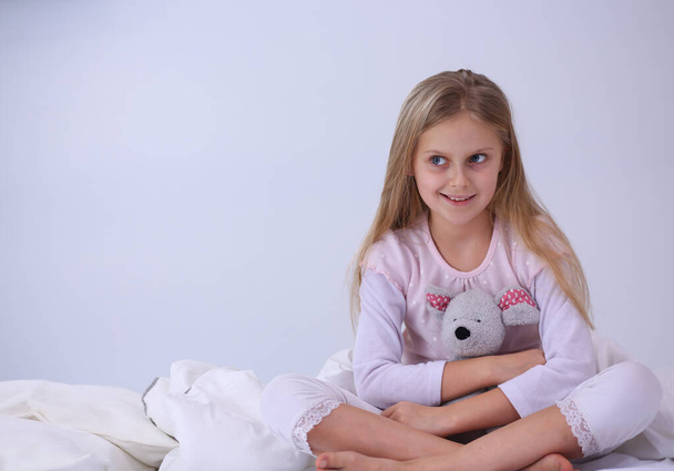Little girl in the bedroom is sitting on the bed. Little girl is wearing a pajamas and sitting in bed . - Foto, immagini