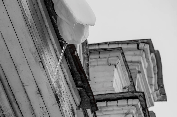Um snowdrift branco com um gelo transparente afiado está pendurado para baixo do telhado cinzento de um tijolo e edifício de madeira no inverno. Preto e branco
 - Foto, Imagem