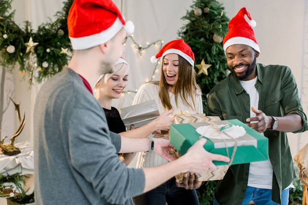 Cheerful young four international people celebrate Christmas and New Year together in cozy decorated studio. Group of friends laughing and sharing Christmas gifts - Foto, Bild