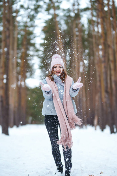 Cute young woman  is playing with snow  in the forest. The girl is dressed in a blue coat, in knitted hat, scarf and mittens. Winter lifestyle happiness emotions nature. Christmas, New year. - Photo, Image