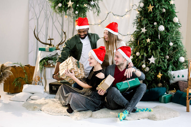 Fiatal, többnemzetiségű barátok csoportja a szépen díszített karácsonyfa közelében, karácsonyi ajándékok cseréje és szórakozás. - Fotó, kép