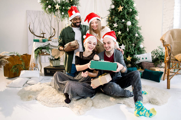 Nouvel An, Noël et vacances d'hiver concept. Jeunes amis multiethniques dans des chapeaux de Père Noël assis sur le sol et célébrant Noël, tenant des boîtes cadeaux et s'amusant
. - Photo, image