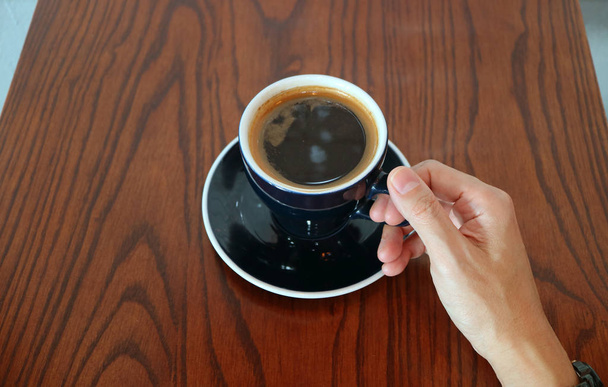 Man Hand Holding de Beker van hete zwarte koffie geserveerd op houten tafel - Foto, afbeelding