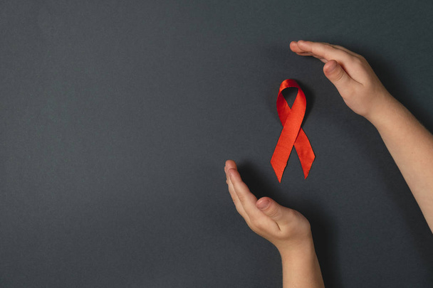 Top view on child's hands holding red ribbon on dark background, HIV awareness concept, world AIDS day. Flat lay, copy space - Foto, imagen