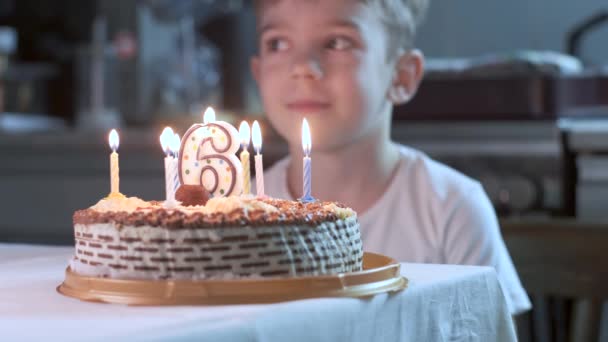 boy sitting at kitchen table and blowing candles on birthday cake, making wish - Materiał filmowy, wideo