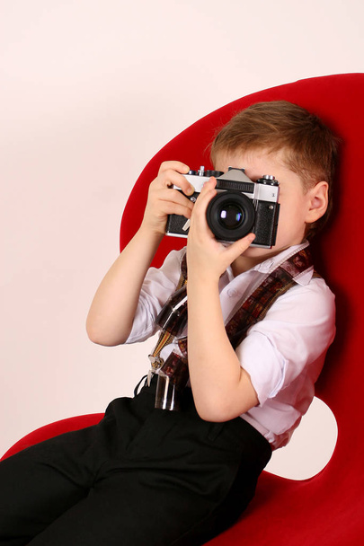 Ragazzino fotografo sedersi in sedia studio rosso con macchina fotografica vintage pellicola e guardare mirino annuncio
 - Foto, immagini