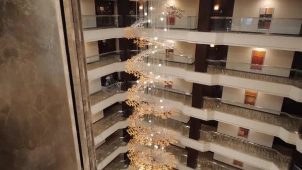 Un hotel urbano caro y hermoso, una lámpara de araña dorada en espiral con ascensor en el centro de la sala. Desde la planta superior hay una vista de un hotel de lujo con muchos pisos y puertas. Movimiento lento
. - Metraje, vídeo