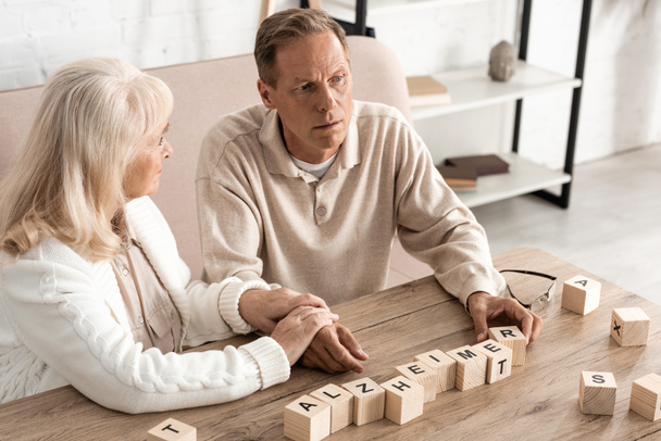 upset senior woman sitting near sick husband near wooden cubes with alzheimer letters  - Photo, Image