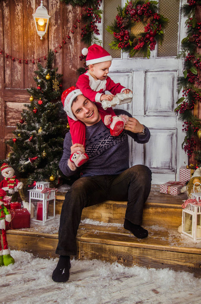 Ευτυχισμένος μπαμπάς στο παιχνίδι καπέλο Σάντα με το μικρό γιο του ντυμένο ως Άγιος Βασίλης στο παρασκήνιο της Πρωτοχρονιάς και των Χριστουγέννων εσωτερικό. - Φωτογραφία, εικόνα