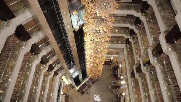 Gezellige en brede lobby van een duur hotel van de bovenste verdieping tot de eerste. Twee liften van het hotel gaan naar beneden. Onderaan bij de receptie zitten mensen. Langzame beweging. - Video