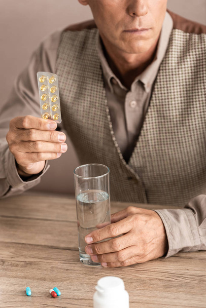 обрізаний вид на пенсіонера з хворобою Альцгеймера, що тримає таблетки і склянку води
  - Фото, зображення