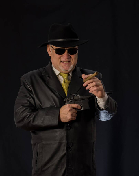  A man in black clothes, a black leather coat, hat. Ganster with a cigar Mauser system pistol. Mafiosi in black glasses on a dark background. Severe, strong, stylish, impressive, dangerous, unfriendly, - Foto, Imagen