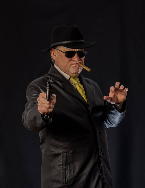 Ένας άντρας με μαύρα ρούχα, μαύρο δερμάτινο παλτό, καπέλο. Ganster με ένα πούρο σύστημα Mauser πιστόλι. Μαφιόζι με μαύρα γυαλιά σε σκούρο φόντο. Σοβαρή, δυνατή, κομψή, εντυπωσιακή, επικίνδυνη, εχθρική, - Φωτογραφία, εικόνα