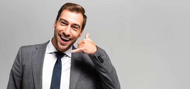πανοραμική φωτογραφία του χαμογελαστού επιχειρηματία με κοστούμι δείχνει σήμα κλήση απομονώνονται σε γκρι  - Φωτογραφία, εικόνα