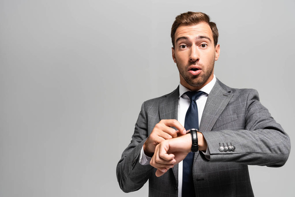 όμορφος και σοκαρισμένος επιχειρηματίας με κοστούμι με ρολόι χειρός κοιτάζοντας την κάμερα που απομονώνεται σε γκρι  - Φωτογραφία, εικόνα