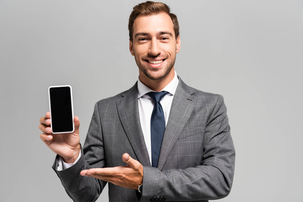 pohledný a usměvavý podnikatel v obleku ukazuje rukou na smartphone izolované na šedé  - Fotografie, Obrázek