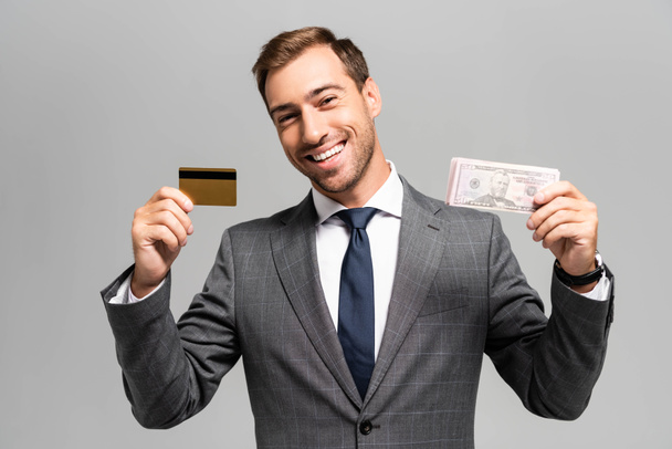 handsome and smiling businessman in suit holding credit card and dollar banknotes isolated on grey  - Zdjęcie, obraz