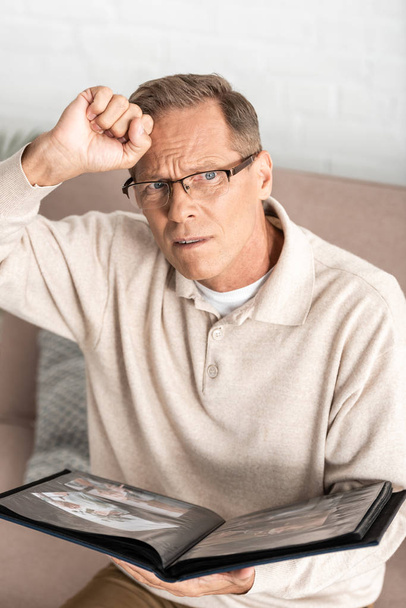 zdenerwowany starszy mężczyzna w okularach trzyma album fotograficzny  - Zdjęcie, obraz