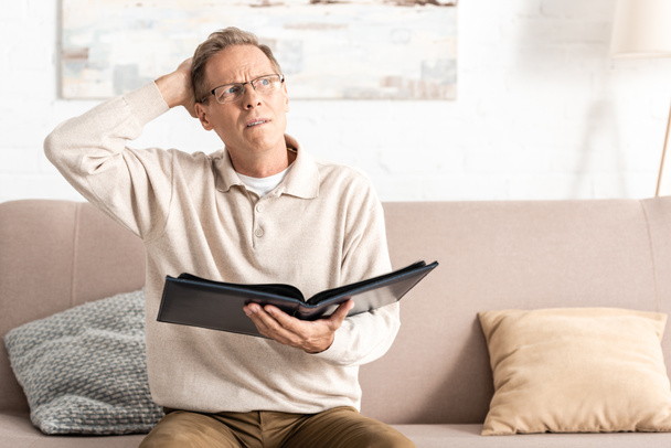confused senior man in glasses holding photo album while sitting on sofa  - Photo, Image