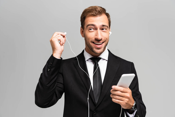 handsome and smiling businessman in suit with earphones using smartphone isolated on grey  - Foto, afbeelding