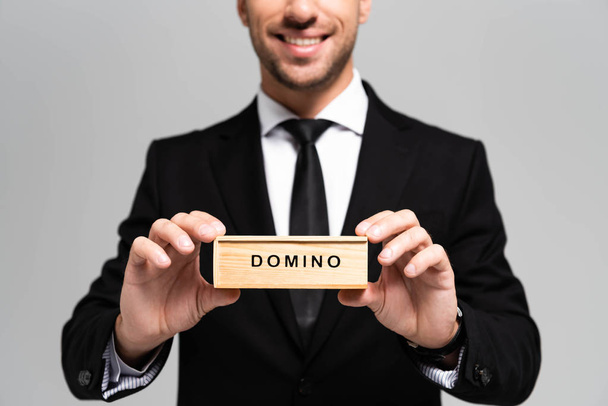 обрізаний вид усміхненого бізнесмена в костюмі, що тримає дерев'яну коробку з написом доміно ізольовано на сірому
  - Фото, зображення