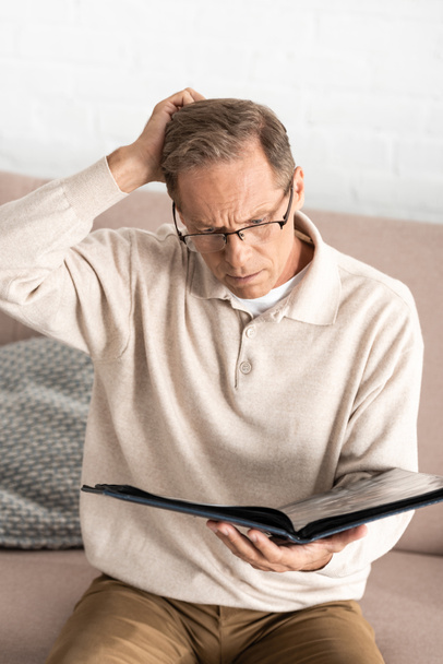 στοχαστικός άνδρας με γυαλιά με νόσο Alzheimer κρατώντας άλμπουμ φωτογραφιών στο σπίτι  - Φωτογραφία, εικόνα