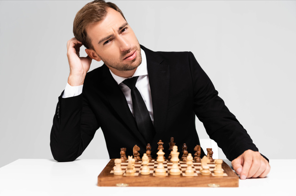pensive and handsome businessman in suit sitting near chessboard isolated on grey  - Фото, изображение