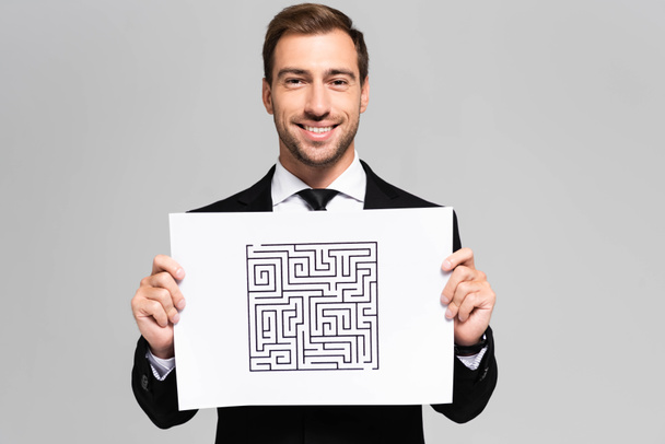 usmívající se a pohledný obchodník držící papír s labyrintem izolovaným na šedi  - Fotografie, Obrázek