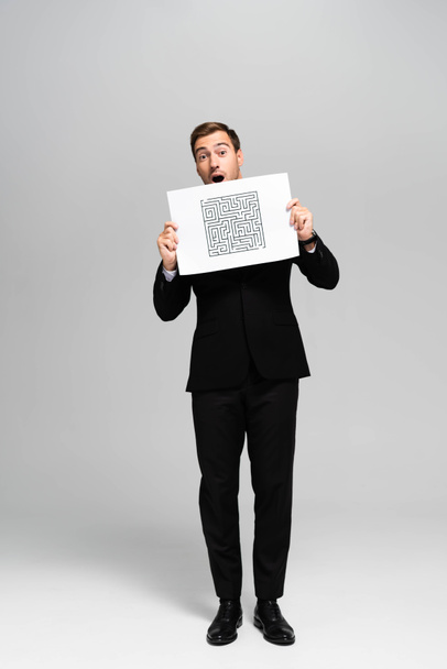 όμορφος και σοκαρισμένος επιχειρηματίας με κοστούμι κρατώντας χαρτί με λαβύρινθο σε γκρι φόντο  - Φωτογραφία, εικόνα