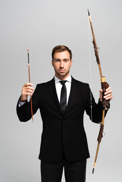 handsome businessman in suit holding bow and arrow isolated on grey  - 写真・画像