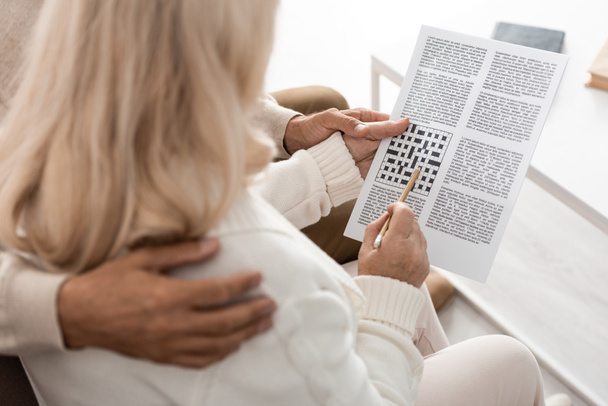 cropped view of senior man hugging wife with pencil and crossword  - Photo, Image
