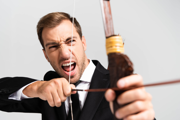 selective focus of angry businessman in suit holding bow and shooting with arrow isolated on grey - Foto, imagen