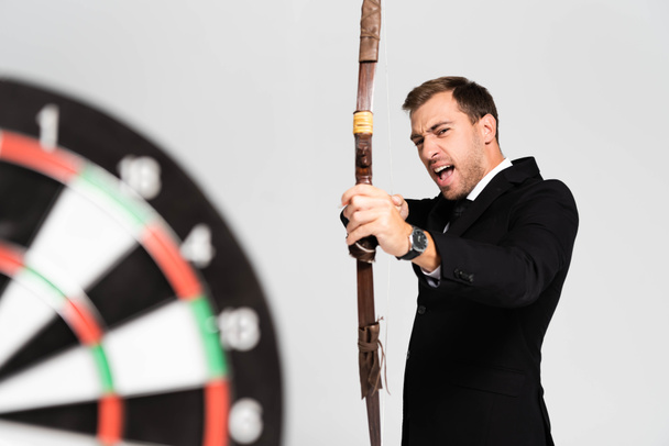 selective focus of angry businessman in suit holding bow and shooting at target isolated on grey - 写真・画像