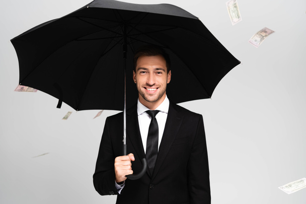 przystojny i uśmiechnięty biznesmen w garniturze trzymający parasol i stojący blisko spadających banknotów dolarowych odizolowanych na szaro - Zdjęcie, obraz