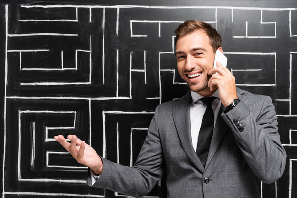hombre de negocios guapo y sonriente en traje hablando en el teléfono inteligente y de pie cerca del laberinto
 - Foto, imagen