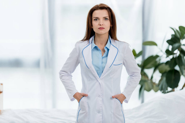 attractive doctor in white coat with hands in pockets looking at camera in hospital  - Фото, зображення